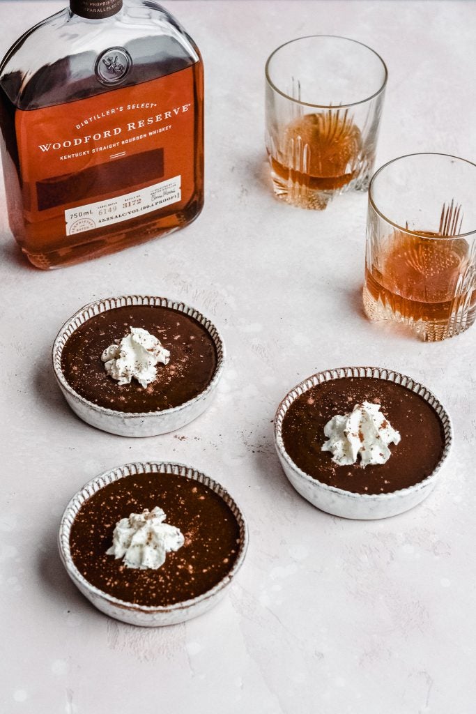 Three Chocolate Bourbon Pots de Creme on a white counter with two neat glasses of Woodford Reserve Kentucky Straight Bourbon Whiskey and an open bottle of Woodford Reserve Kentucky Straight Bourbon Whiskey